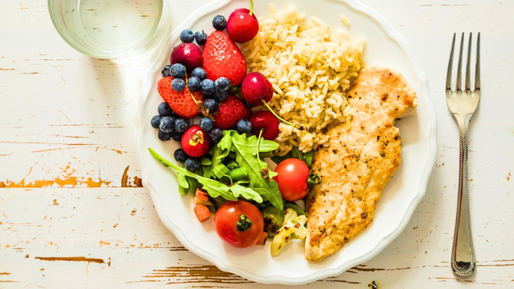 A plate of food including a piece of grilled chicken breast, a serving of rice, assorted fresh berries (strawberries, blueberries, and cherries), a small salad with lettuce, cherry tomatoes, and avocado, and a fork beside the plate. A glass of water is in the background.