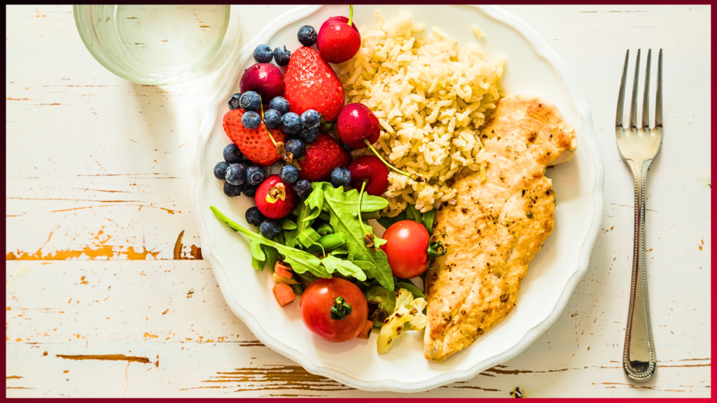 A white plate with a meal consisting of grilled chicken breast, a serving of brown rice, and a side salad featuring tomatoes, mixed greens, and a mix of strawberries, raspberries, cherries, and blueberries. A glass of water and a fork are placed beside the plate.