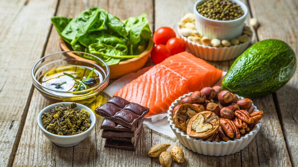 A wooden table is filled with a variety of nutritious foods, including a salmon fillet, sliced avocado, spinach, cherry tomatoes, nuts, dark chocolate, dried herbs, olive oil, and assorted seeds in bowls.