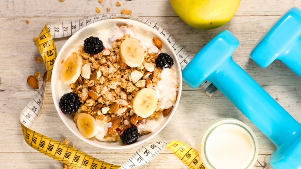 A bowl of granola topped with banana slices, blackberries, and nuts sits on a wooden table next to measuring tape, a green apple, blue hand weights, and a small glass of milk.