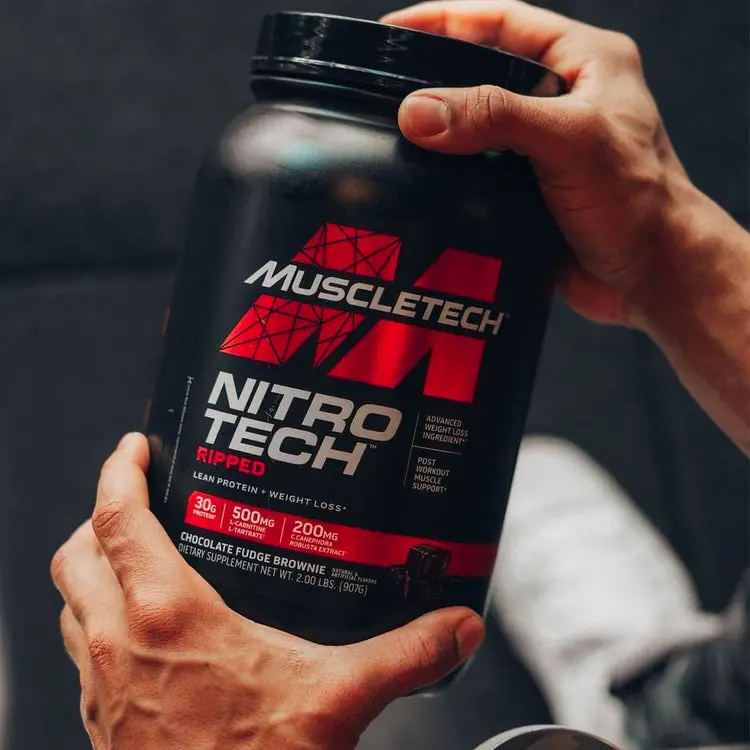 A person holding a black container of MuscleTech Nitro Tech Ripped protein powder with a red label. The container states "Chocolate Fudge Brownie" flavor and highlights 30g of protein, 500mg of CLA, and 200mg of C. Canephora Robusta. Hands are seen gripping the container.