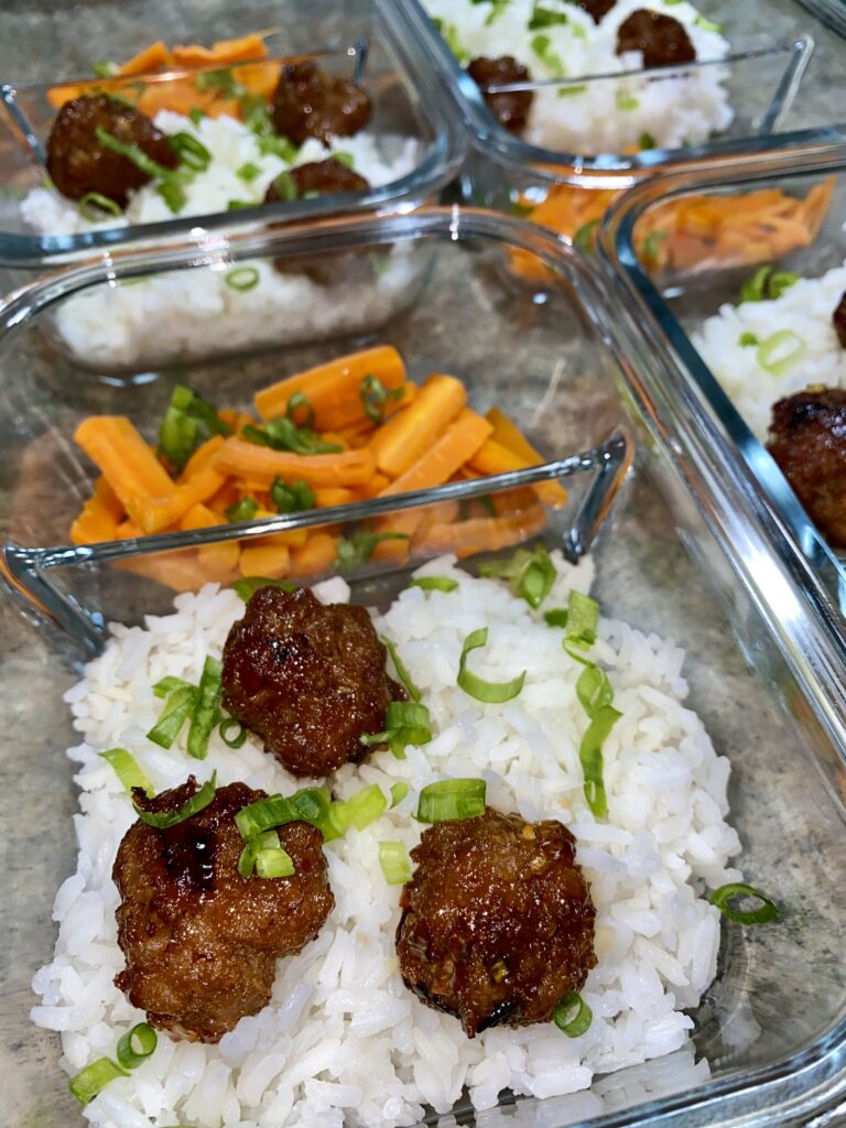 Glass containers filled with white rice topped with small, saucy meatballs and sprinkled with chopped green onions. Adjacent compartments contain julienned carrots. The containers are arranged on a gray countertop.