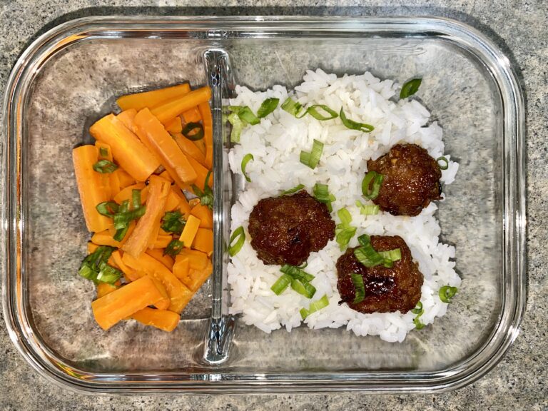 A meal in a glass container with a compartment divider. One side has sliced orange carrots topped with chopped green onions. The other side has white rice, three meatballs, and is garnished with chopped green onions.