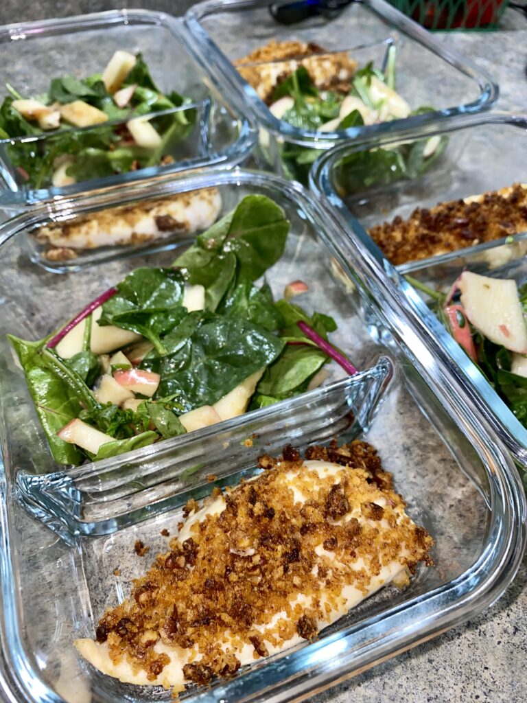 Four glass meal prep containers on a countertop, each containing a portion of breaded baked chicken and fresh spinach salad with apple slices. The meal is divided evenly in separate sections within the containers.