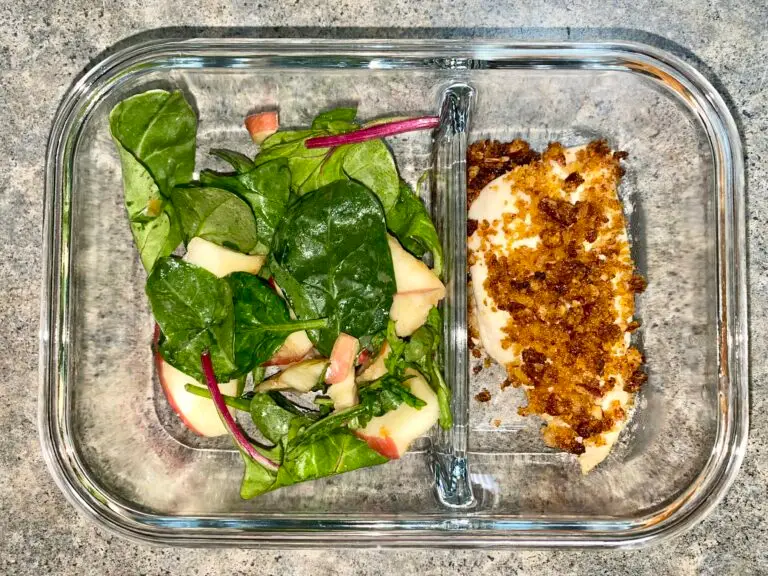 A divided glass meal prep container holds a fresh green salad with apple slices in the left compartment and a serving of baked chicken topped with breadcrumbs in the right compartment, placed on a gray countertop.