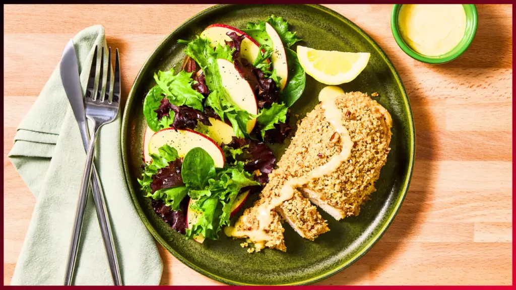 A plate with a breaded chicken breast garnished with a lemon wedge, accompanied by a mixed greens salad with apple slices. A small green bowl of dressing is visible in the top right corner. Fork and knife rest on a folded green cloth napkin to the left.