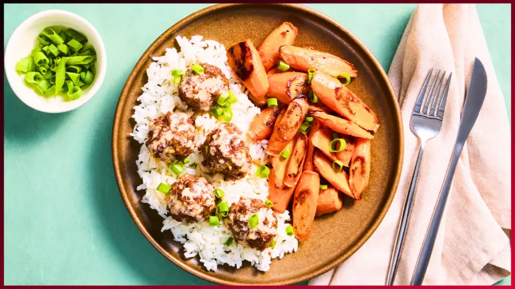 A plate with white rice topped with meatballs garnished with chopped green onions, accompanied by roasted carrot slices. A small bowl to the side contains additional chopped green onions. A knife and fork rest on a folded napkin beside the plate.