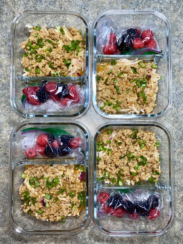 Four glass meal prep containers on a countertop. Each container holds seasoned ground turkey, shredded cheese, and chopped green onions on one side, and a small plastic bag of fresh berries on the other side. The berries are a mix of raspberries and blackberries.