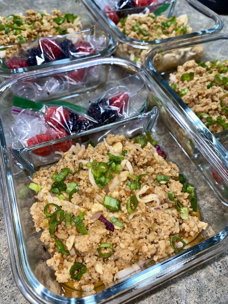 Glass meal prep containers filled with ground turkey seasoned with green onions and paired with bags of mixed fresh berries, including raspberries and blackberries, are neatly arranged on a counter.