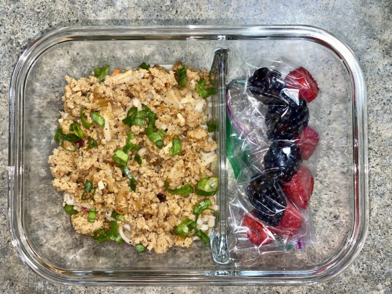 A glass container holds stir-fried ground meat with chopped green onions on the left side. On the right side, there are two small ziplock bags containing blackberries and raspberries, separated from the meat by a glass divider. The background is a speckled countertop.