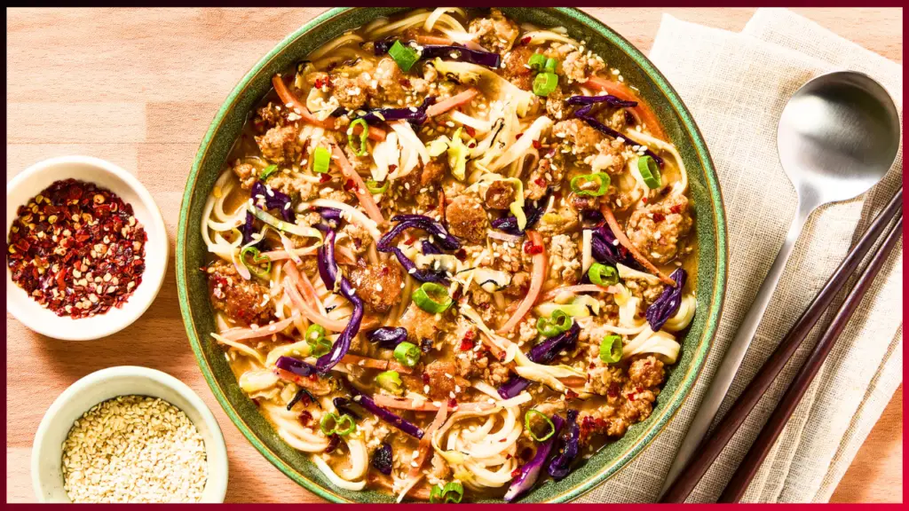 A vibrant bowl of noodle stir-fry features colorful vegetables, ground meat, and garnished with chopped green onions. Accompanying the bowl are small dishes of red chili flakes and sesame seeds. Chopsticks and a spoon rest on a beige linen napkin beside the bowl.