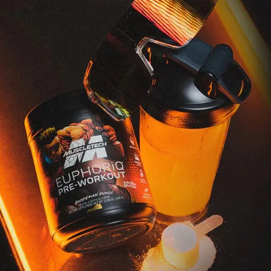 A container of Muscletech EuphorQ pre-workout powder sits next to a filled blender bottle. A scoop and some powder are in front of the container, all placed on an illuminated surface.