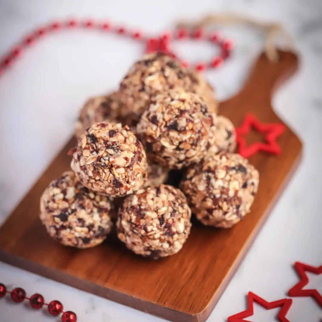 A stack of energy balls with oats and dried fruits on a wooden cutting board, surrounded by festive red star decorations and beads on a marble surface, complements the Holiday Treats [1] - eBook.