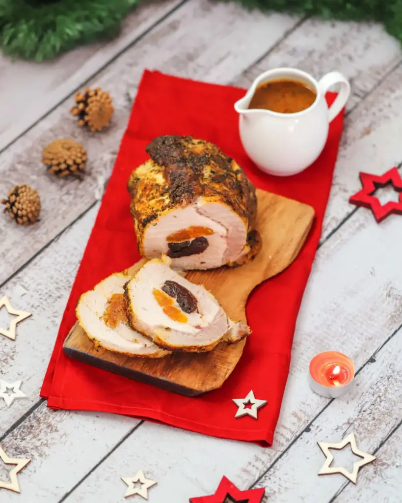 Sliced roast pork loin with a filling, placed on a wooden board over a red napkin. A gravy boat is in the background, along with decorative pine cones, star ornaments, and a small candle on a rustic wooden table.