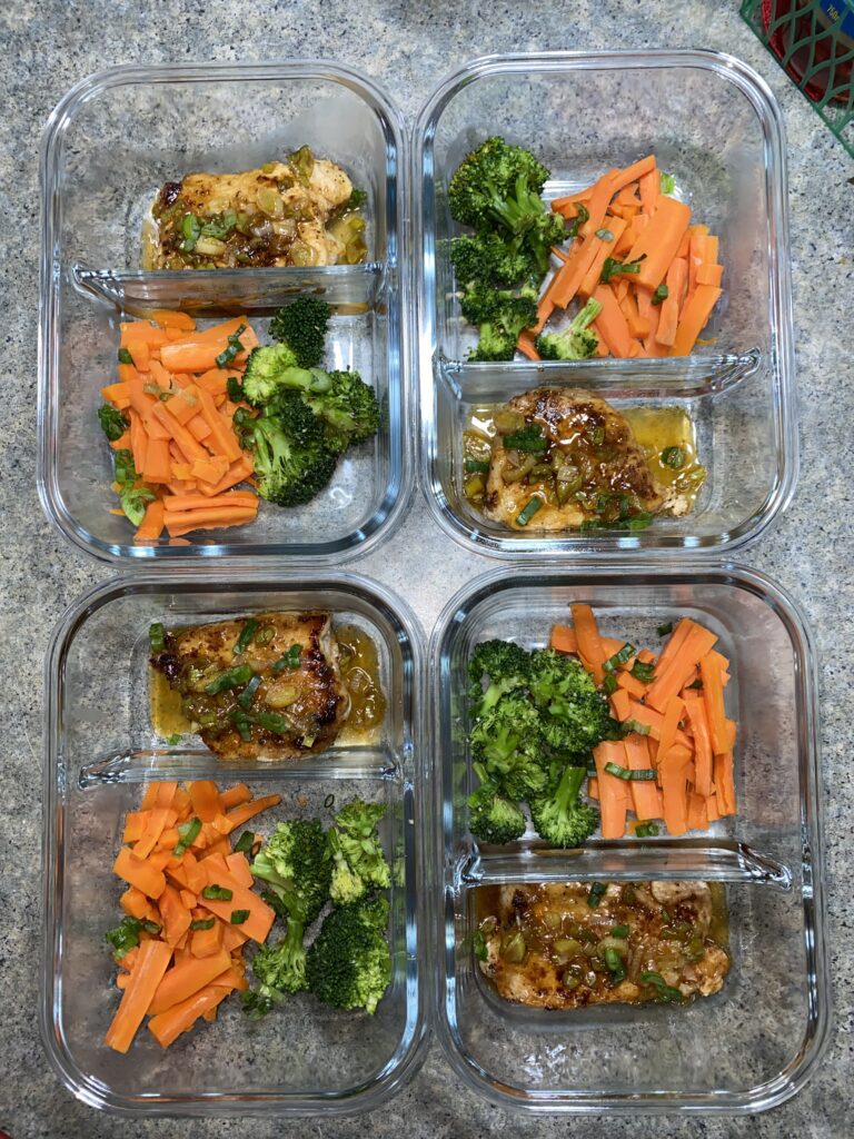 Four glass meal prep containers, each filled with cooked chicken, steamed broccoli, and sliced carrots, arranged neatly on a gray countertop. The chicken is glazed, adding a shiny finish, and garnished with green herbs.