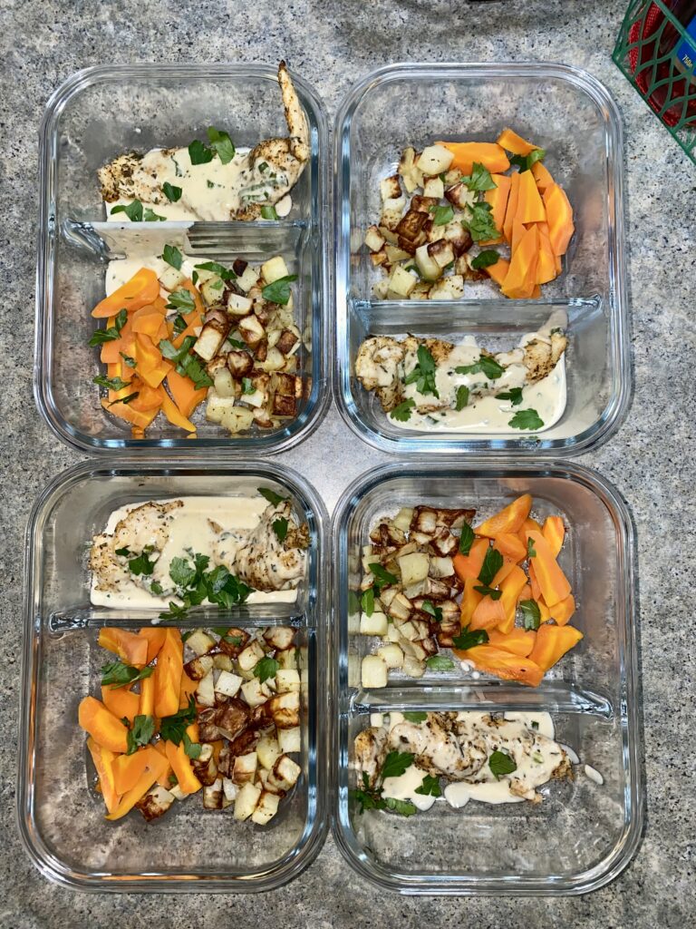 Six glass meal prep containers are filled with roasted chicken, a creamy sauce, cooked potatoes, and sliced carrots, garnished with fresh herbs. They're neatly arranged on a speckled countertop.