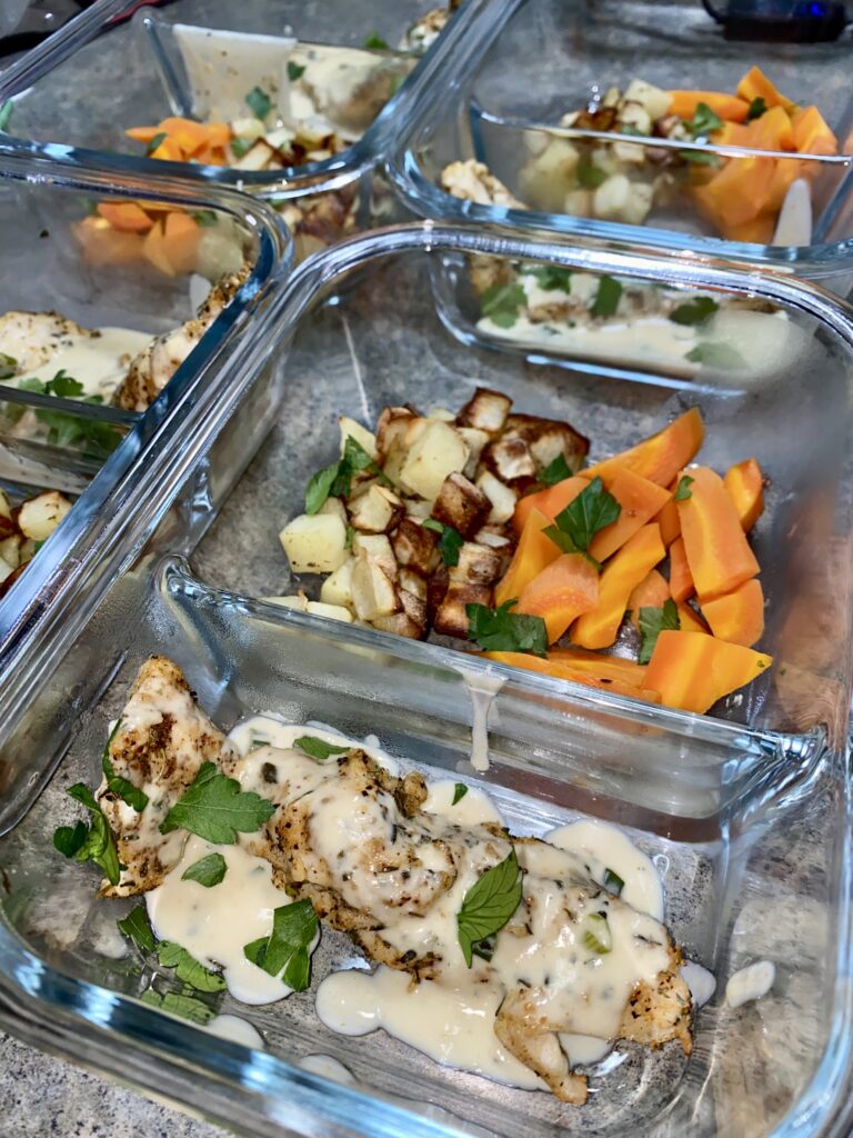 Glass containers filled with meal prep portions of grilled chicken topped with creamy sauce and herbs, roasted potatoes, and carrots are neatly arranged on a kitchen countertop.