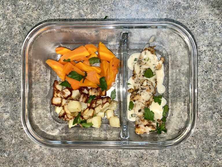 Glass container with baked chicken topped with sauce and herbs on one side, and roasted carrots with parsley, and potatoes on the other. The container is on a gray speckled countertop.