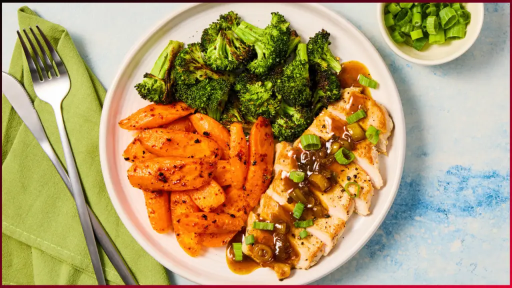 A white plate with sliced pork topped with sauce and green onions, roasted broccoli, and seasoned roasted carrots. A bowl of chopped green onions is on the side. A fork and knife rest on a folded green napkin.