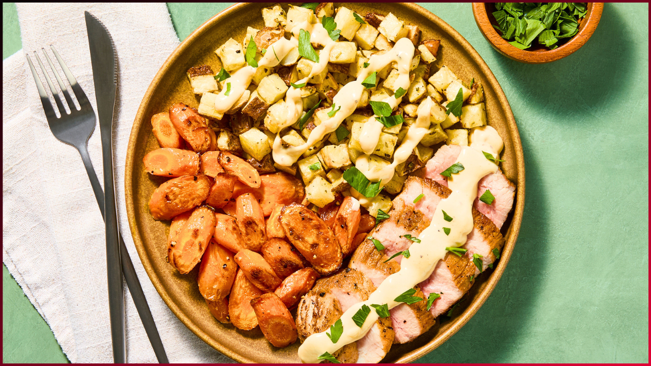 A plate features sliced meat with creamy sauce, roasted diced potatoes, and seasoned carrots, garnished with fresh herbs. Fork and knife rest on a napkin beside the plate, and a small bowl of chopped herbs is nearby.