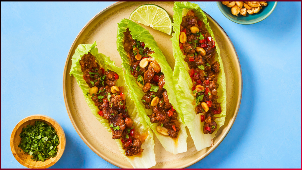 Three lettuce wraps filled with seasoned ground meat, peanuts, and red peppers are arranged on a plate. A lime wedge garnishes the plate. A small bowl of chopped herbs is nearby, all set against a blue background.