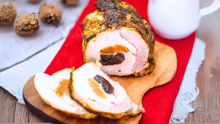 A cooked roulade with slices showing a filling of apricots and prunes is served on a wooden board. Beside it, there's a cream-colored pitcher and truffles. The setup rests on a red and white cloth on a rustic wooden table.