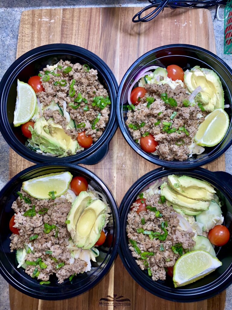 Four black bowls on a wooden surface, each filled with ground meat, sliced avocado, lime wedges, cherry tomatoes, and green onions on a bed of shredded lettuce.