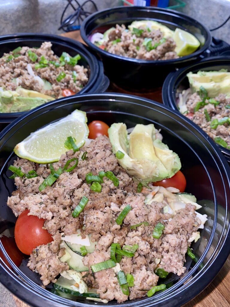 Three black bowls filled with cooked ground meat, avocado slices, cherry tomatoes, shredded cabbage, and topped with chopped green onions. Lime wedges are placed on top for garnish.