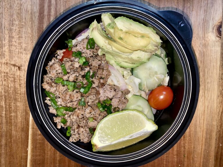 A black bowl on a wooden table filled with ground meat topped with chopped green onions, sliced avocado, cucumber slices, cherry tomatoes, red onions, and a lime wedge.