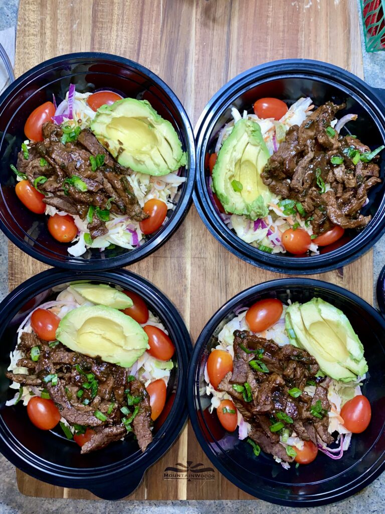 Four bowls of salad with sliced avocado, cherry tomatoes, seasoned beef strips, and shredded cabbage sit on a wooden surface. Each bowl is black and garnished with green herbs.