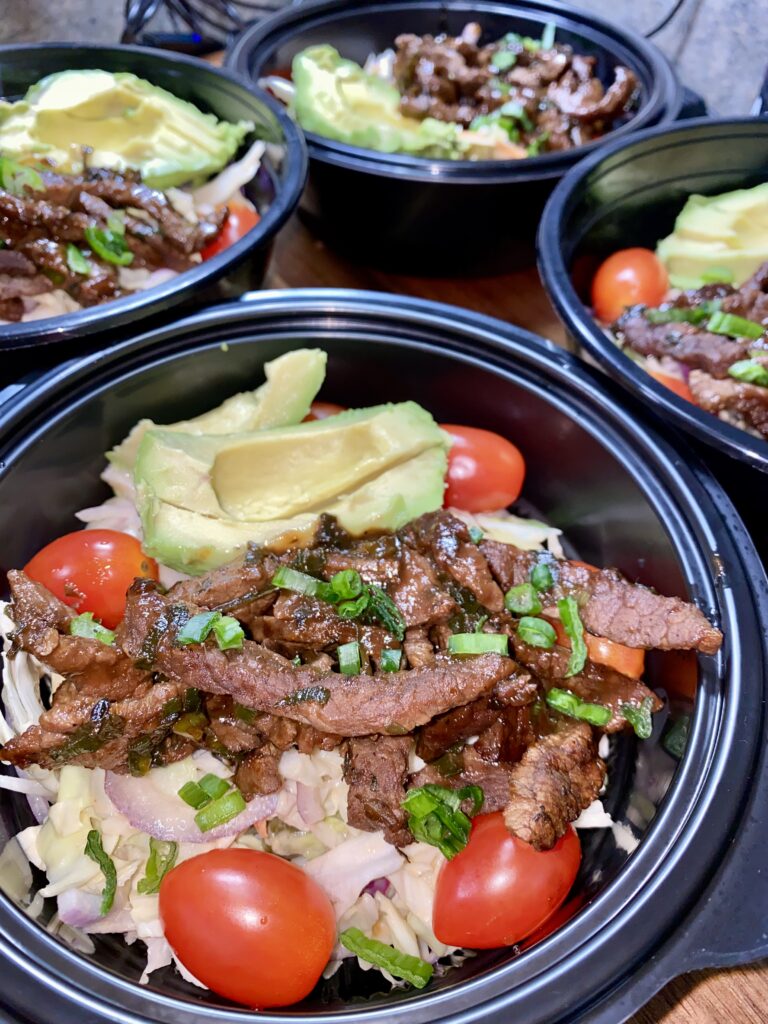 Bowls of salad topped with grilled beef slices, avocado, cherry tomatoes, and chopped green onions. The salad mix includes cabbage and onions, served in black bowls.