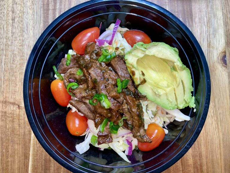 A black bowl contains sliced beef topped with green onions, nestled on a bed of shredded cabbage, cherry tomatoes, and red onion. A half avocado is placed on the side. The bowl is on a wooden surface.