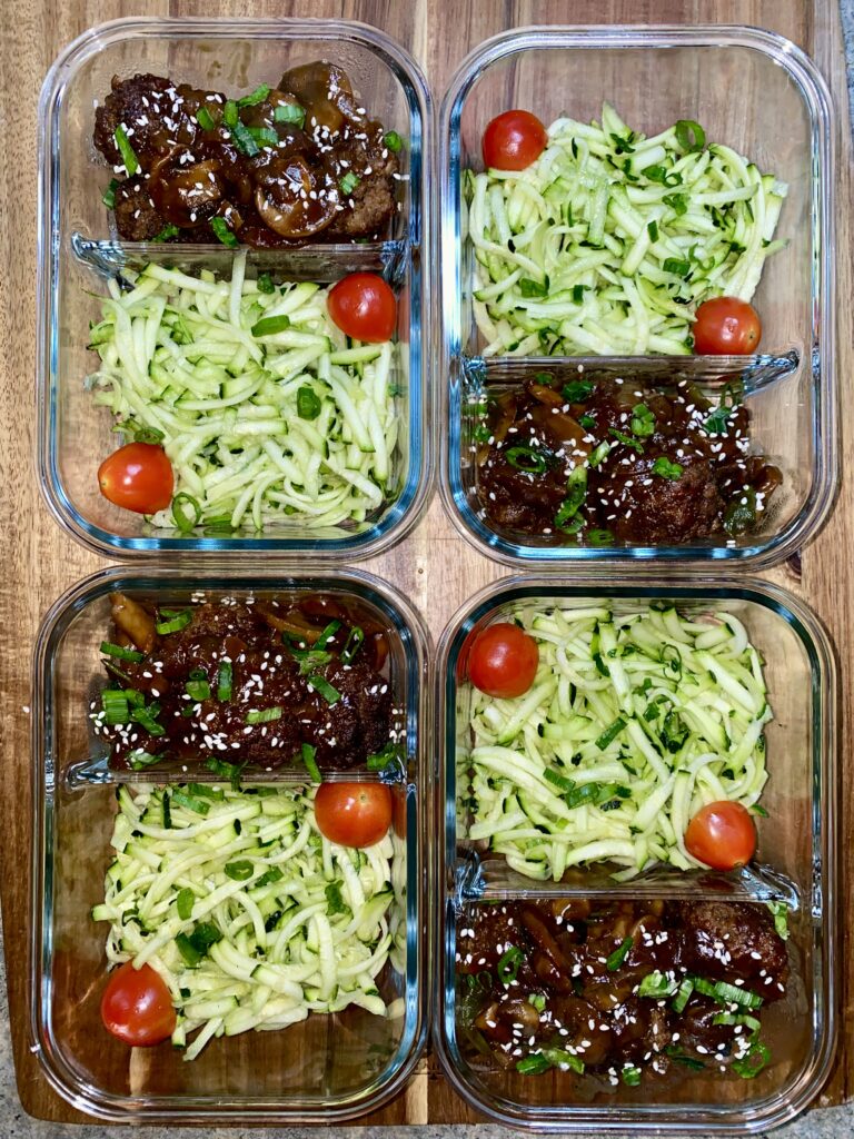 Four meal prep containers are arranged on a table. Each contains sliced zucchini and cherry tomatoes on one side, and marinated beef garnished with sesame seeds and herbs on the other. The containers are placed in a neat grid.