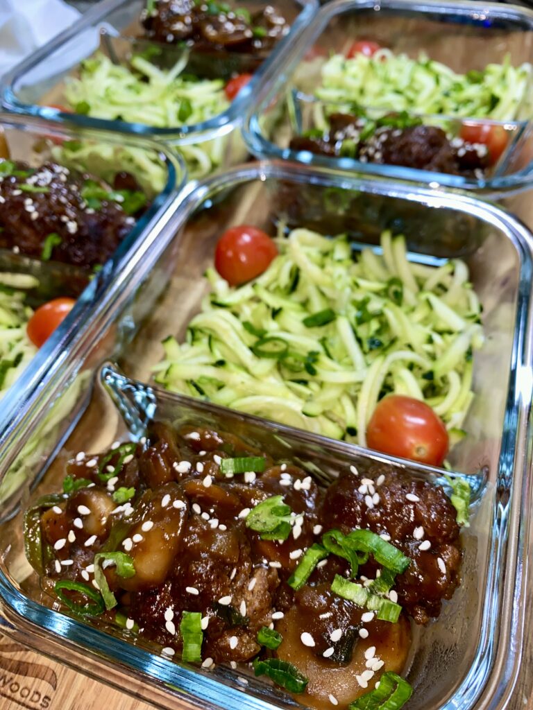 Glass containers filled with sliced beef topped with sesame seeds and green onions, placed alongside zucchini noodles and cherry tomatoes. The meal prep containers are arranged on a wooden surface.