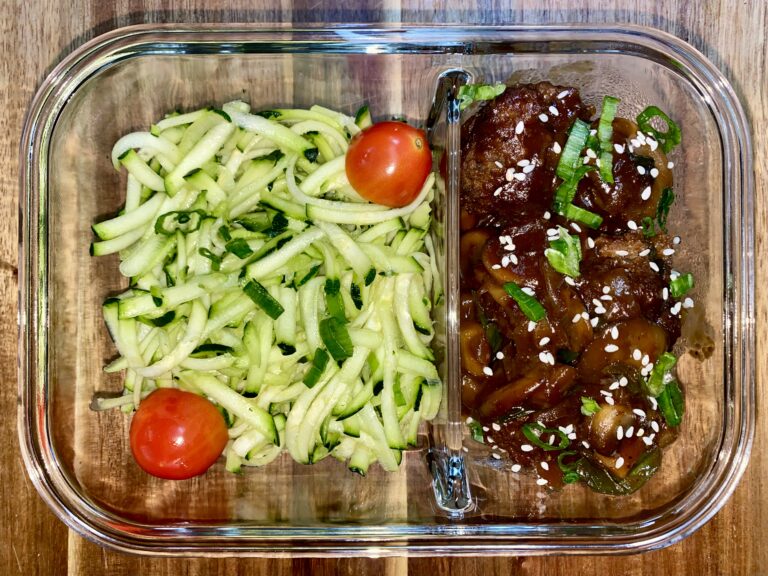 A glass meal prep container with spiralized zucchini and two cherry tomatoes on the left. The right side contains meatballs garnished with sesame seeds and chopped green herbs. The container is placed on a wooden surface.