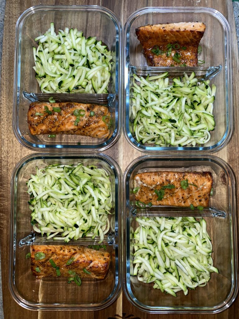 Four glass meal prep containers on a wooden surface, each containing grilled salmon and shredded zucchini noodles. The salmon is garnished with herbs.