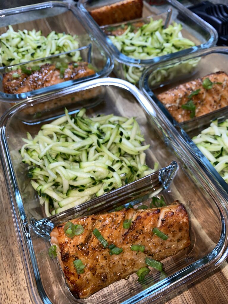 Glass meal prep containers with grilled salmon and shredded zucchini garnish. The salmon is topped with chopped herbs, and the containers are arranged on a wooden surface.