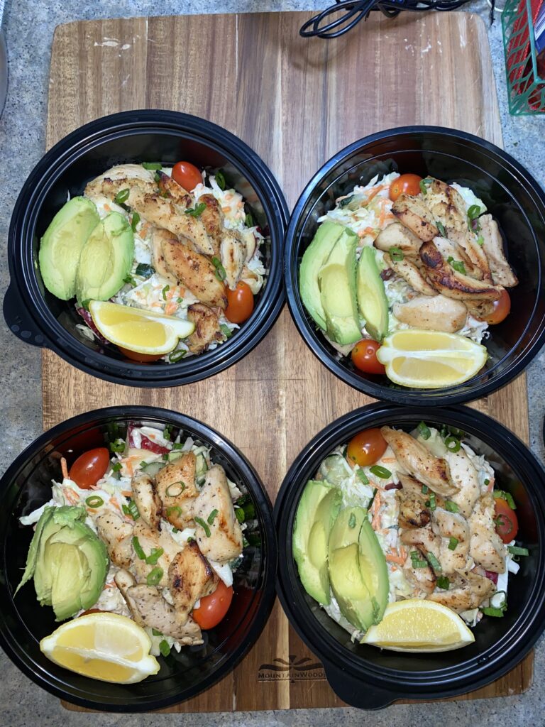 Four black bowls on a wooden surface, each filled with grilled chicken, avocado slices, cherry tomatoes, shredded vegetables, and lemon wedges.