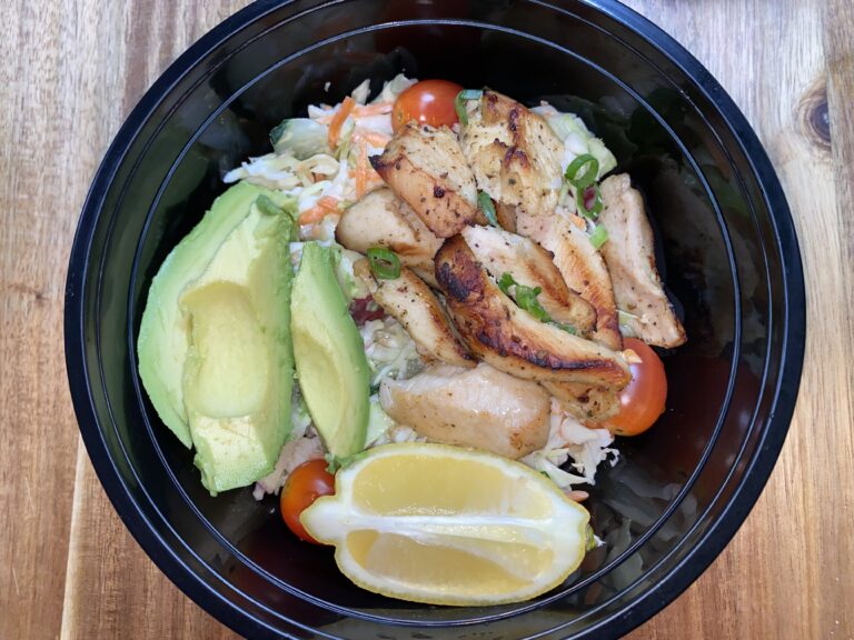 A black bowl filled with a salad topped with grilled chicken slices, avocado, cherry tomatoes, and a lemon wedge on the side. The salad sits on a wooden table.