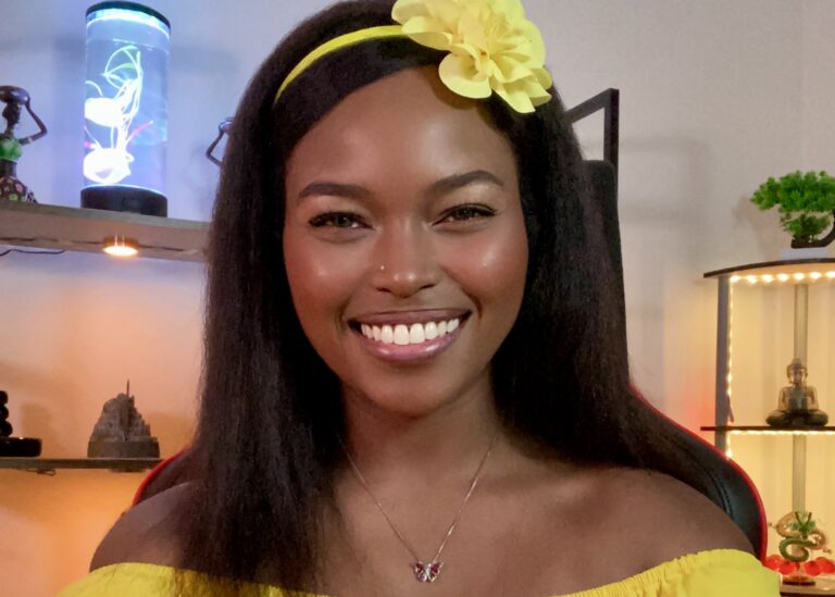 A woman with long dark hair and a yellow flower headband smiles at the camera. She wears a yellow off-shoulder top and a butterfly necklace, with decorative shelves and warm lighting in the background.