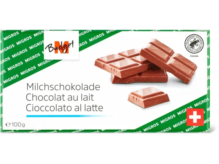 A wrapped bar of milk chocolate labeled “Milchschokolade / Chocolat au lait / Cioccolato al latte” with the Migros Budget logo and a Swiss flag on the packaging. The package is green and white with images of chocolate pieces.