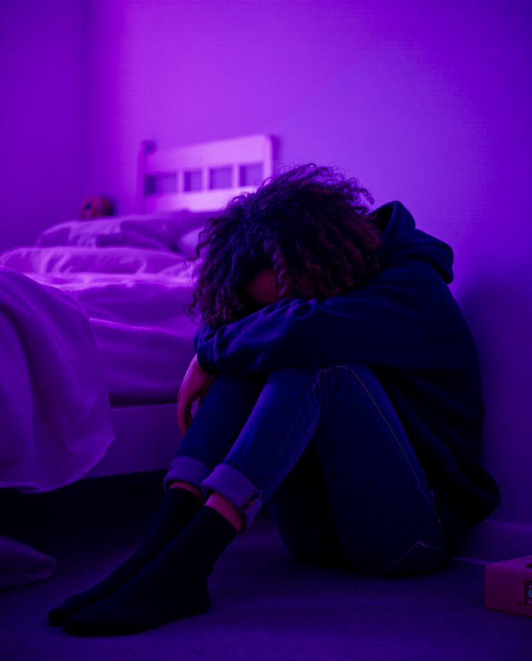 A person sits on the floor by a bed, hugging their knees and resting their head on their arms, in a dimly lit room with purple lighting, creating a somber mood.