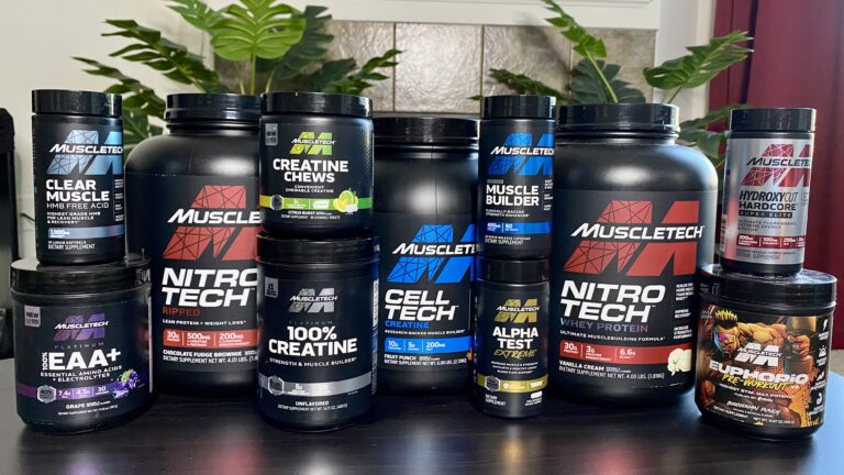 A collection of various MuscleTech supplement containers, including protein powders, creatine, amino acids, and fat burners, displayed on a table with green plants in the background.