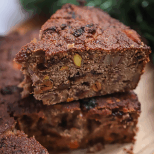 Two thick slices from The Ultimate Holiday Recipe Bundle’s fruitcake, stacked on a wooden surface, display a dense texture with visible nuts and pieces of dried fruit.