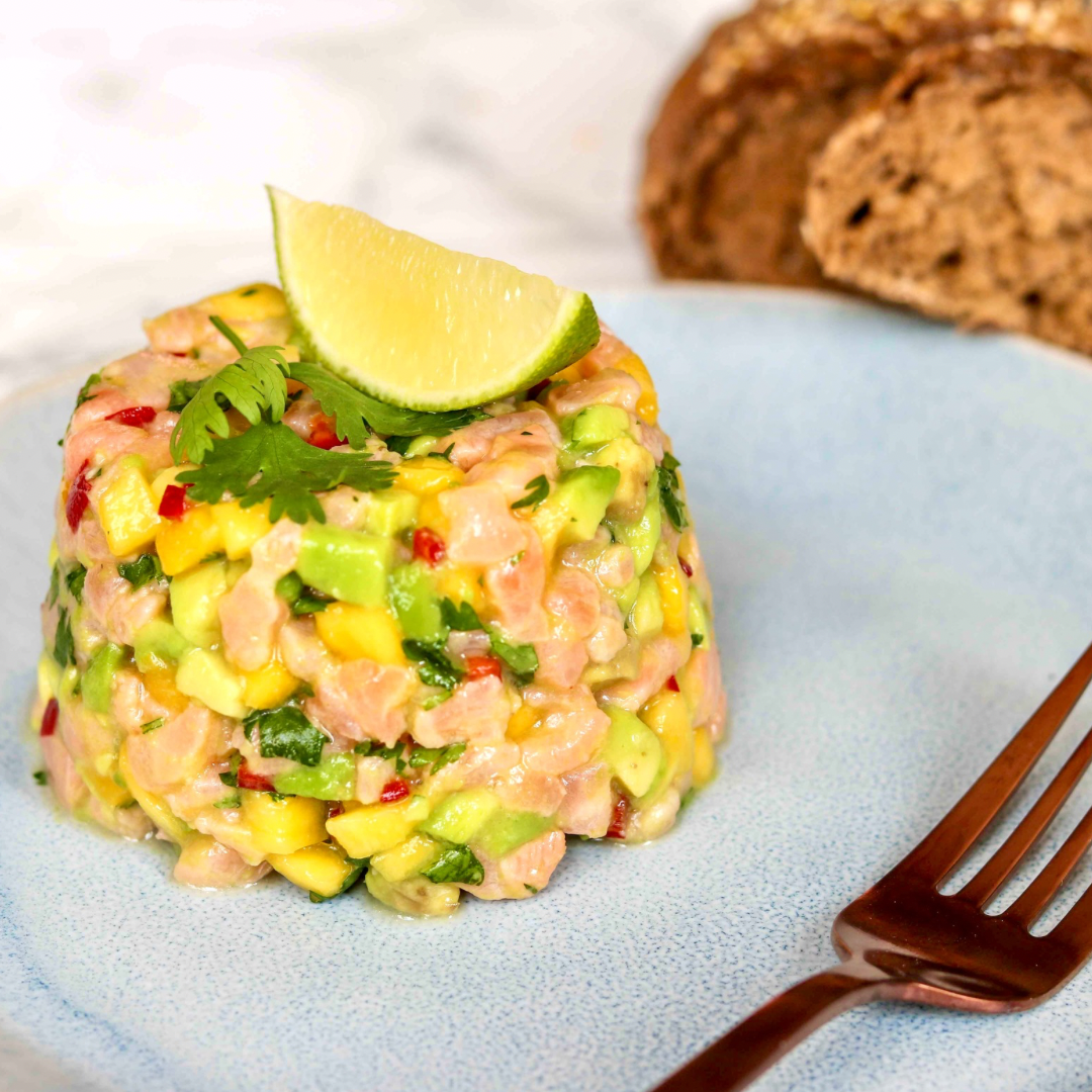 A neatly shaped serving of salmon tartare with avocado, mango, and herbs sits on a blue plate for The New Year Reset Recipe Bundle. A lime wedge tops the dish, while whole grain bread slices appear blurred in the background.