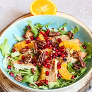 Enjoy a vibrant salad from The New Year Reset Recipe Bundle: grilled chicken slices, orange segments, pecans, pomegranate seeds, and mixed greens with a half orange garnish, all beautifully served on a wooden board.