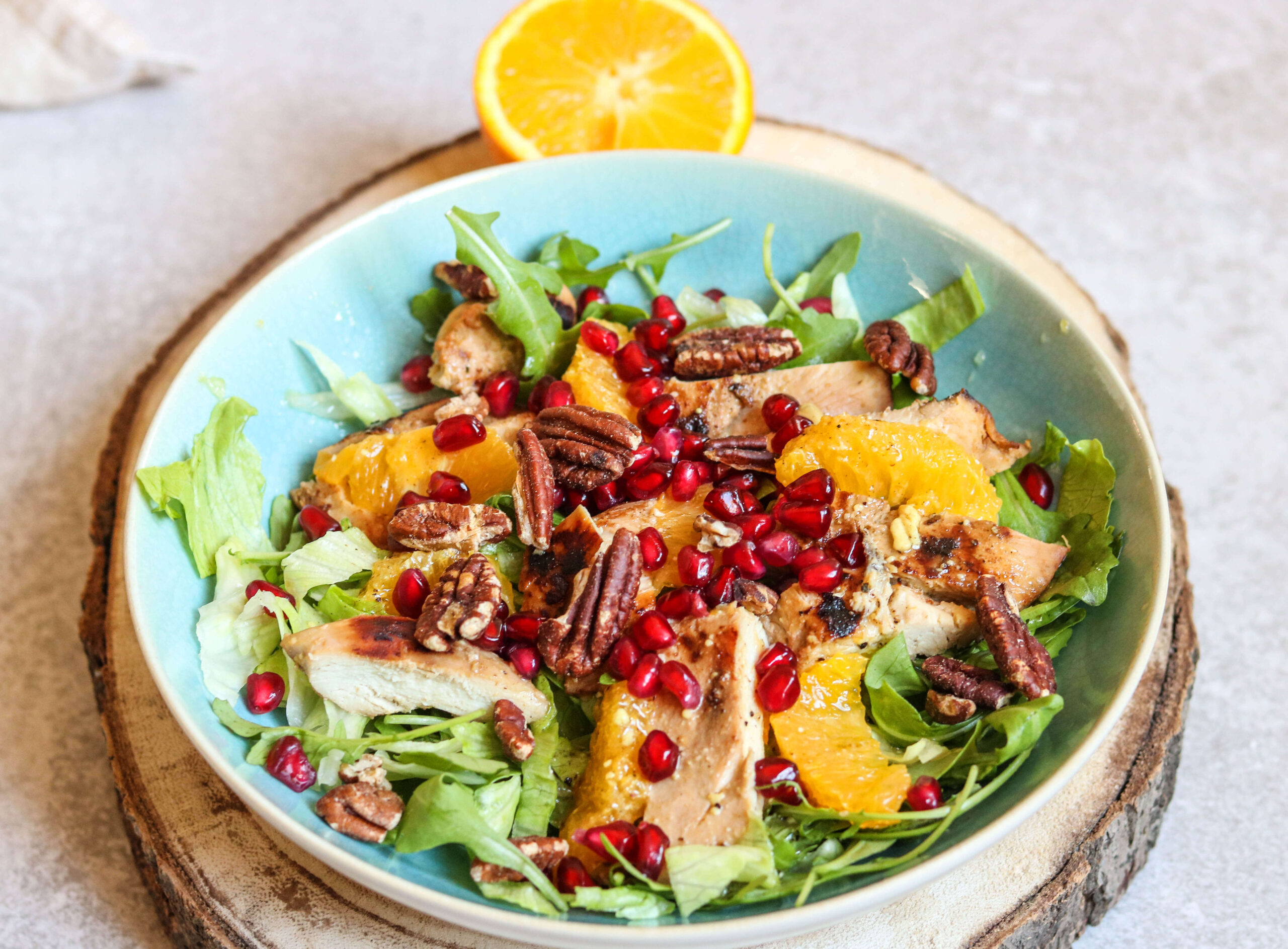 Enjoy a vibrant salad from The New Year Reset Recipe Bundle: grilled chicken slices, orange segments, pecans, pomegranate seeds, and mixed greens with a half orange garnish, all beautifully served on a wooden board.