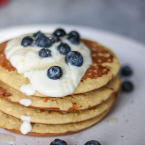 A delicious stack of pancakes from The New Year Reset Recipe Bundle, topped with creamy yogurt and fresh blueberries, served on a white plate with extra blueberries scattered around.