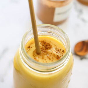 A glass jar filled with a creamy yellow smoothie from The New Year Reset Recipe Bundle, topped with cinnamon and a straw. In the blurred background, a jar of cinnamon powder and a spoon can be seen.