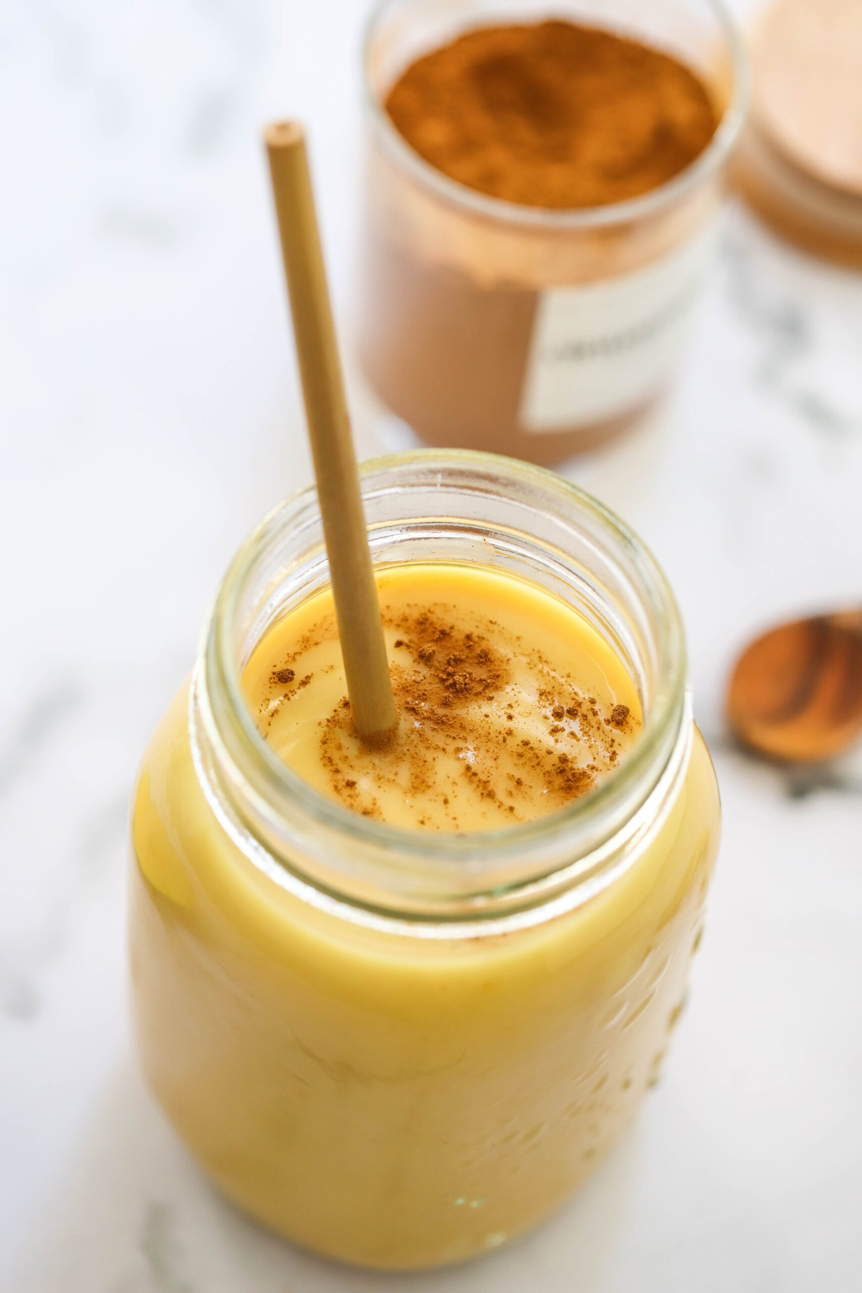 A glass jar filled with a creamy yellow smoothie from The New Year Reset Recipe Bundle, topped with cinnamon and a straw. In the blurred background, a jar of cinnamon powder and a spoon can be seen.